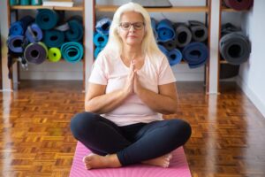 Senior woman meditating on yoga mat, promoting relaxation and wellness indoors.