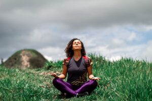A woman enjoys peaceful meditation in a grassy field under a cloudy sky in Brazil.