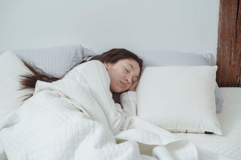 From above of young calm female with long dark wavy hair in white nightwear sleeping on soft pillow in bed under blanket in morning at home